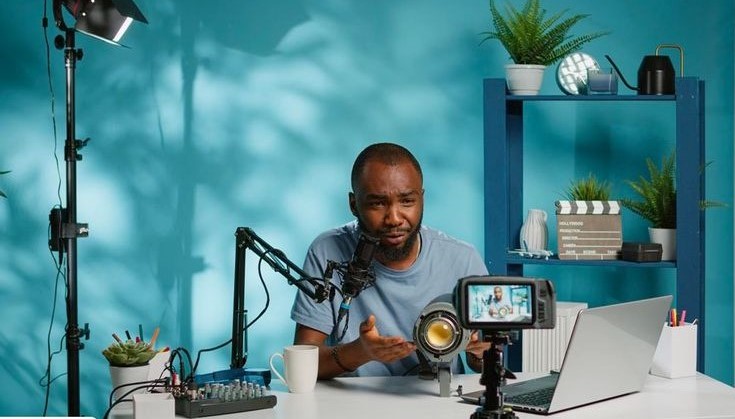A man seated in a blue-colored room with recording apparatus on the table in front of him.